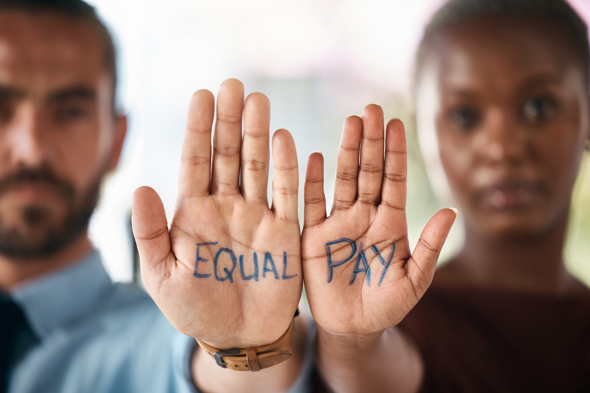 A white man and a black woman hold their palms to the camera. "Equal Pay" is written on their palms in marker