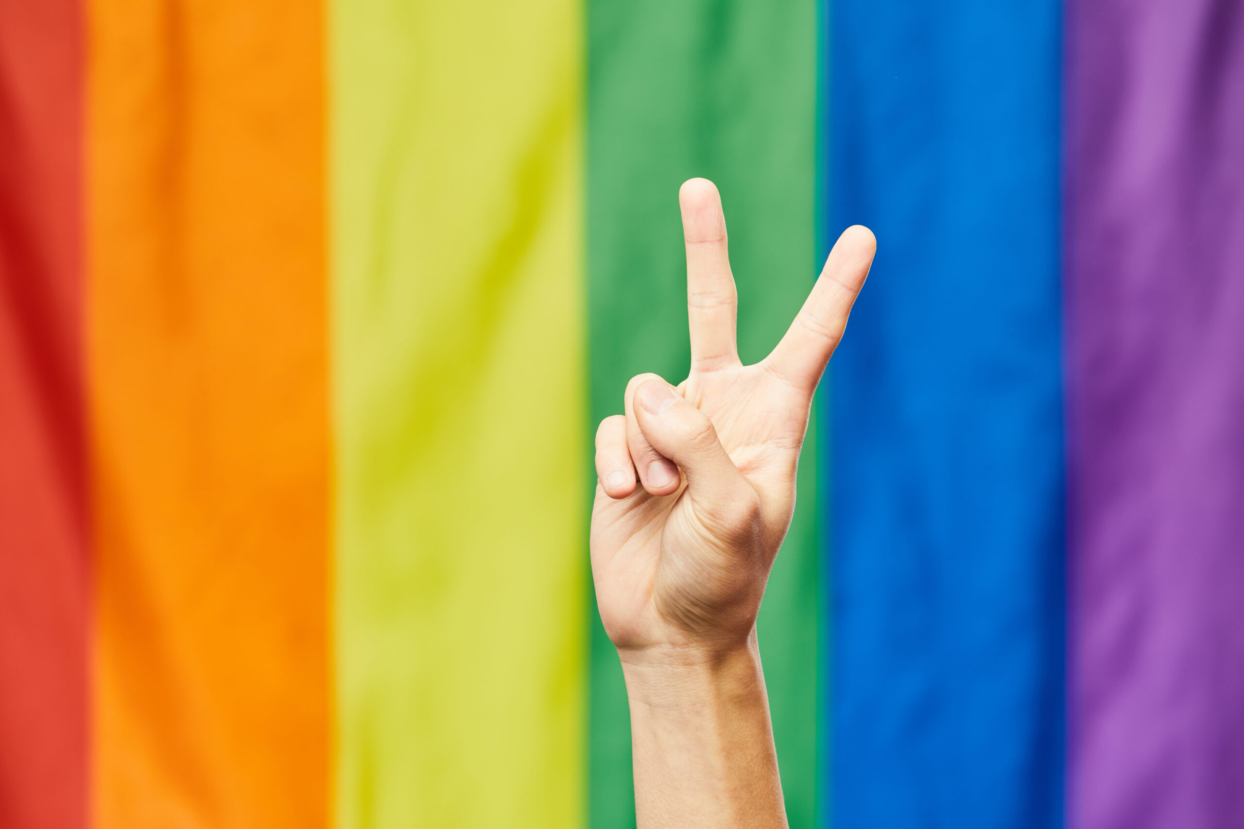 a hand makes a peace sign in front of an LGBTQ+ flag